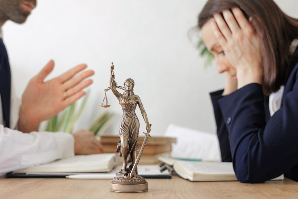 A stressed woman at a table with a Lady Justice statue sits across from a person in a white button-up shirt.