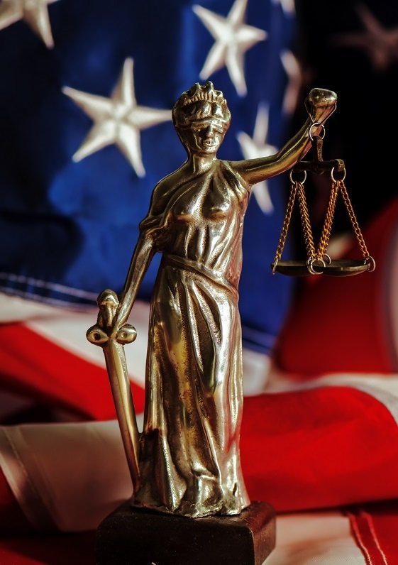 A statuette of Lady Justice holding a sword and scales in front of an American flag.