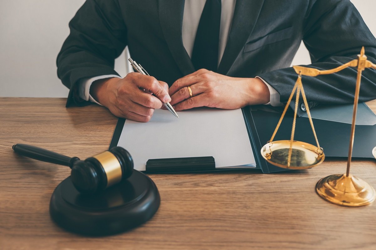 A lawyer writing on a document on a desk next to a gavel and set of scales.
