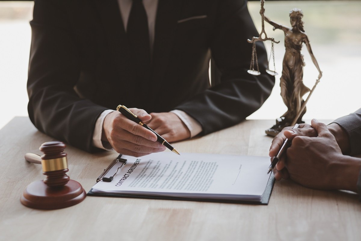 An attorney pointing with a pen at a contract in front of a client at a table with a gavel and Lady Justice.