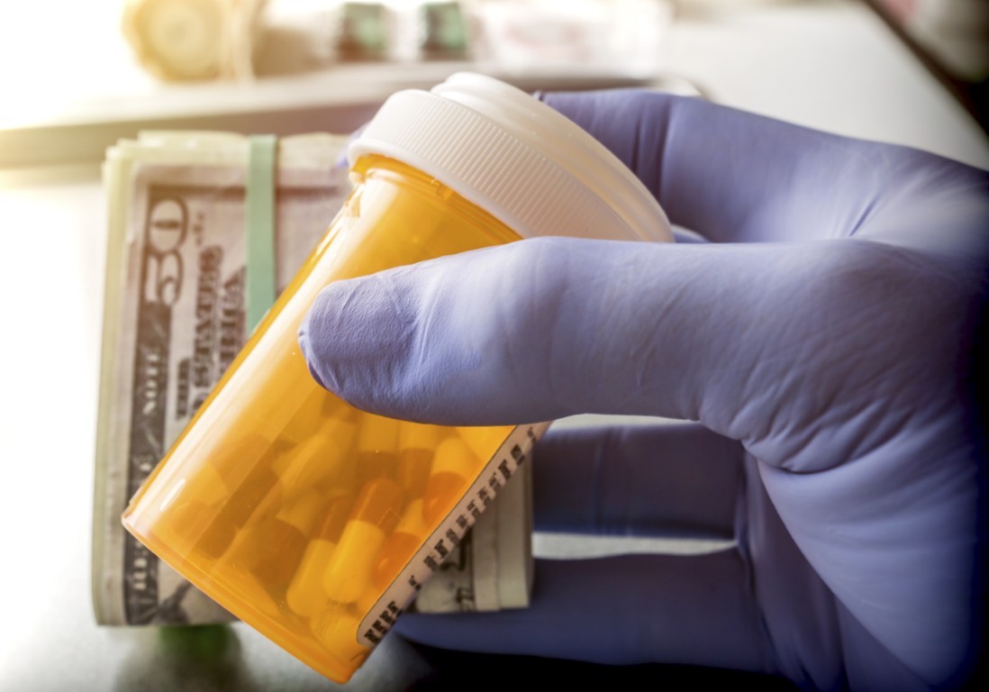 A person in rubber gloves passing a bottle of pills and wad of money to an unseen person